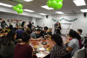 People eating at tables with green balloons. Large Nubian Message poster on wall in background.