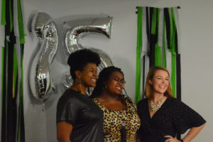 Three women pose in front of silver balloons and green streamers at photo booth